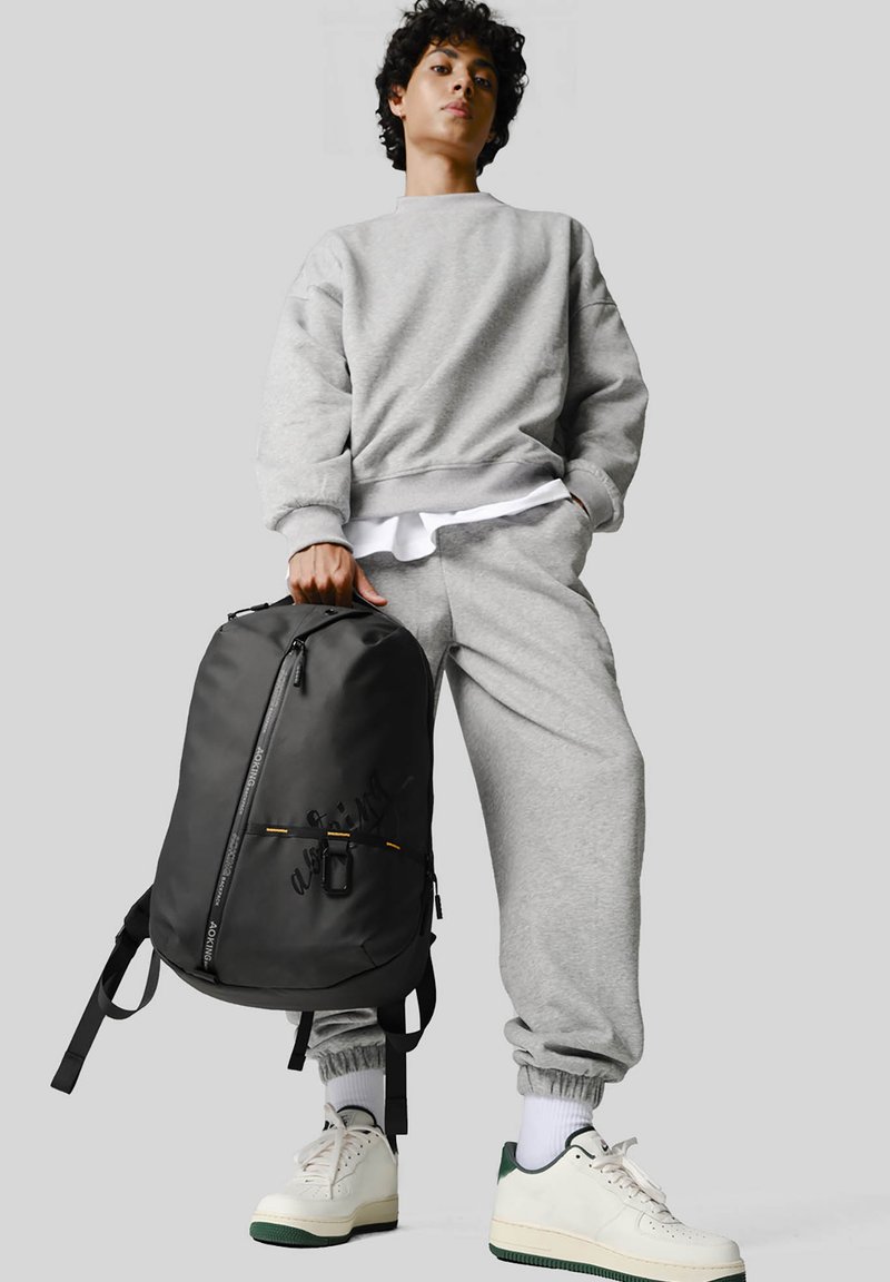Sweat-shirt gris et pantalon de jogging avec des poignets élastiques, associés à un sac à dos noir avec des accents jaunes et un zip avant. Baskets blanches.