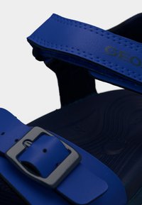 Niebieska sandał z regulowanym paskiem z klamrą i rzepem przy kostce, z teksturowaną wkładką i częściową nazwą marki na pasku górnym.