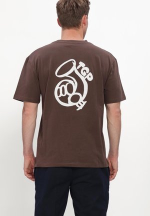 Mann trägt braunes T-Shirt mit weißem abstraktem Horn- und Buchstabendesign auf dem Rücken, kombiniert mit dunkelblauer Hose.