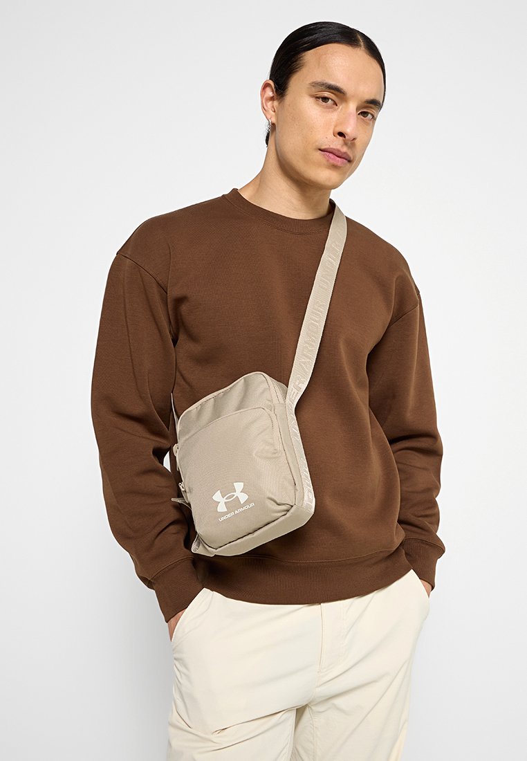 Sac à bandoulière beige en tissu texturé, avec une sangle ajustable et un logo Under Armour blanc ; porté sur un sweat-shirt marron.