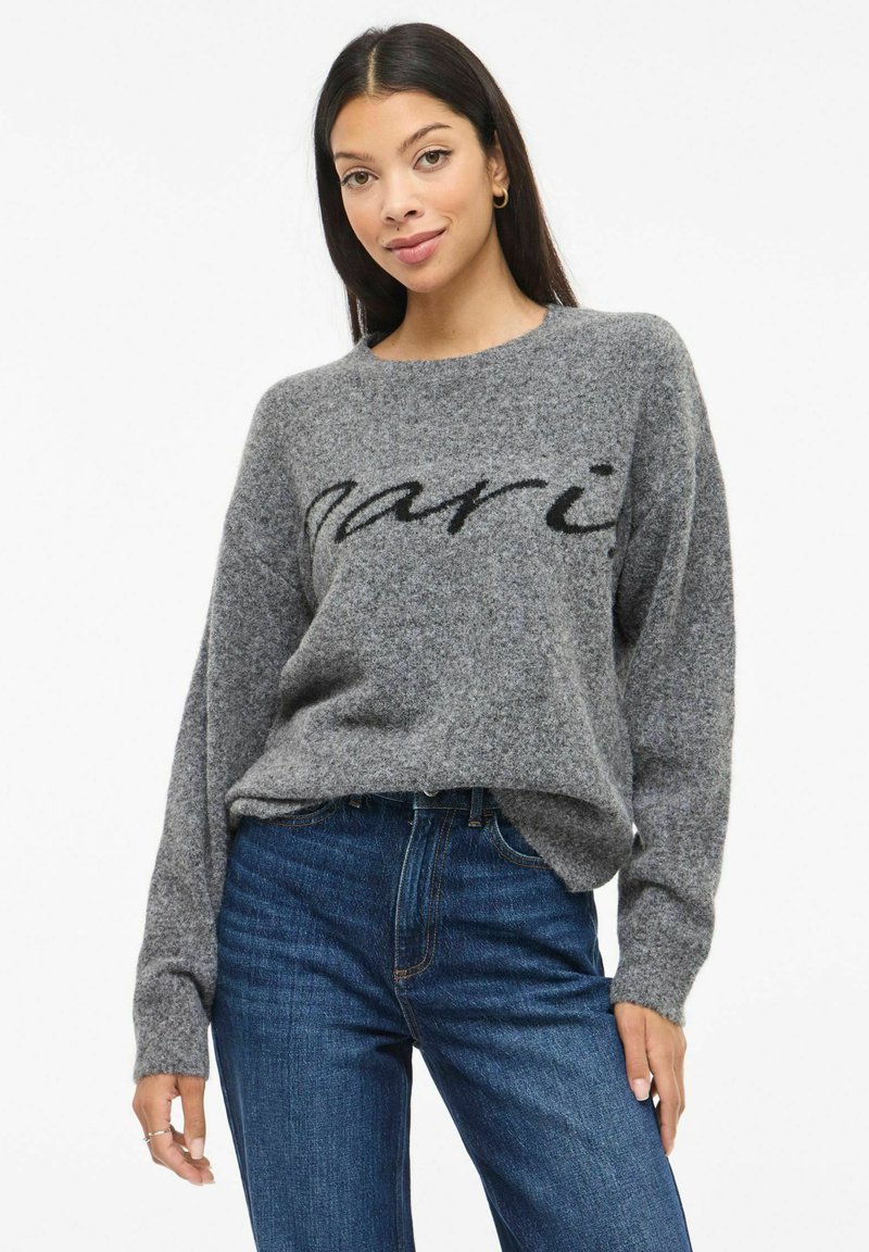 Suéter gris extragrande con una textura suave, con la palabra "Paris" en negro en el frente, combinado con jeans azules. Mangas largas.