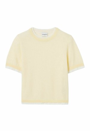 Donna con capelli lunghi e scuri che indossa una maglietta a maniche corte di colore giallo pallido, pantaloni larghi blu con motivo, scarpe marroni e una piccola borsa bianca a tracolla.