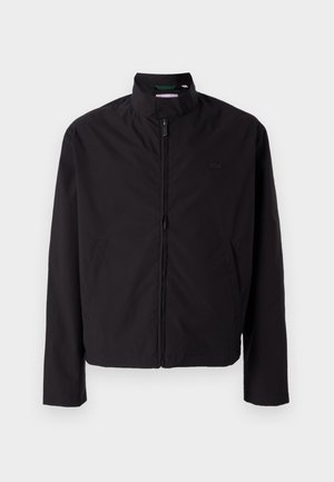 Schwarze leichte Jacke, mit Reißverschluss, Stehkragen, Seitentaschen, glattes Material, minimalistisches Design mit Logo-Akzent auf der Brust.