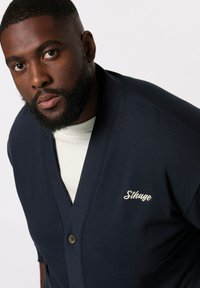 Cárdigan azul marino hecho de una tela suave, con un escote en V profundo, un cierre de un solo botón y el logotipo bordado "Sthuge" en el lado izquierdo.