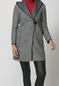 Cappotto a scacchi grigio e nero con ampio colletto, chiusura a bottoni frontale e due tasche laterali; abbinato a leggings neri.