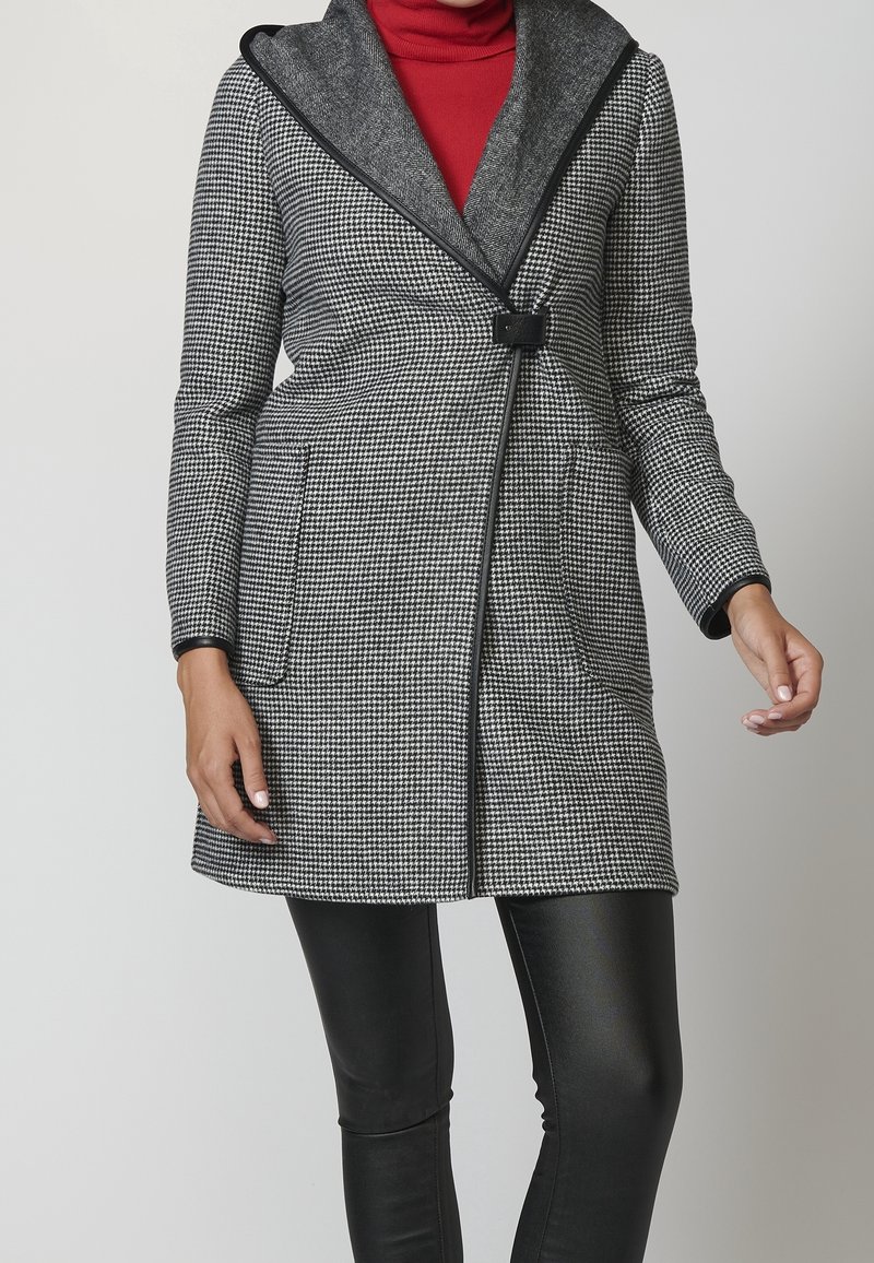 Cappotto a scacchi grigio e nero con ampio colletto, chiusura a bottoni frontale e due tasche laterali; abbinato a leggings neri.