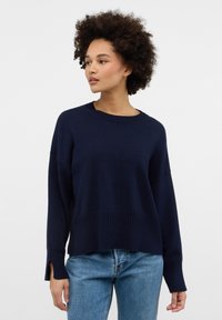 Marineblauer Strickpullover mit gerippten Akzenten und seitlichen Schlitzen; lockere Passform und Rundhalsausschnitt, kombiniert mit hellblauen Jeans.