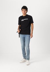 Fekete pamut póló "KARL LAGERFELD JEANS" fehér logóval, világoskék farmernadrággal és fehér sportcipővel párosítva. Álló póz.