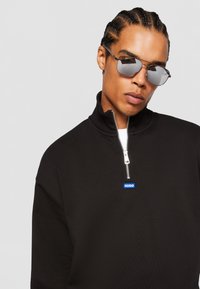 Black zip-up sweatshirt with high collar, featuring a silver zipper and a blue logo patch. Model is wearing mirrored sunglasses.