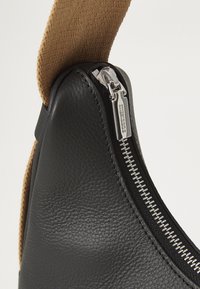 Borsa in pelle nera con finitura testurizzata, dotata di chiusura con zip in argento e una tracolla intrecciata color sabbia, che evidenzia un design funzionale e moderno.