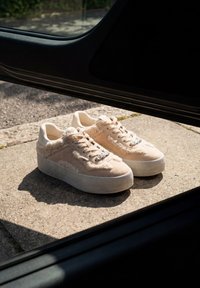 Baskets à plateforme beige avec des détails duveteux posées sur un sol en béton devant une porte de voiture ouverte en pleine lumière du jour.