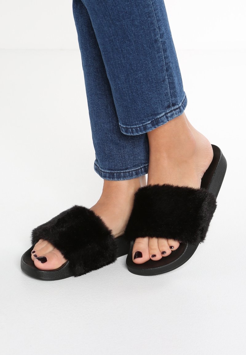Pieds portant des sandales noires en peluche associées à un jean bleu foncé sur un fond blanc uni.