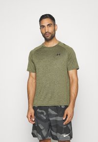 Olive green short-sleeve athletic shirt with a textured finish and black logo on the chest, paired with dark patterned shorts.