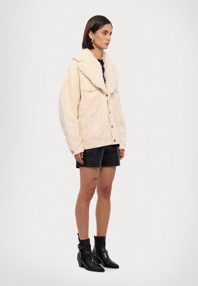 Chaqueta oversized color crema con un cuello esponjoso, múltiples bolsillos frontales y cierre de botones. Usada sobre unos pantalones cortos negros y botas tobilleras.