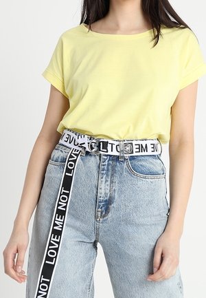 Camiseta amarilla de manga corta combinada con jeans de mezclilla azul de cintura alta. El cinturón presenta un atrevido estampado en blanco y negro de "NO ME AMES".