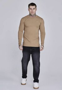 Duga majica s okruglim izrezom, uparena s tamnim trapericama i bijelim tenisicama. Na prsima se nalazi diskretan logotip.