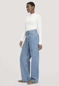 Luokat leveät siniset farkut korkealla vyötäröllä ja edessä solmittavalla yksityiskohdalla, yhdistettynä istuvaan valkoiseen pitkähihaiseen puseroon. Sileä pinta ja rento tyyli.