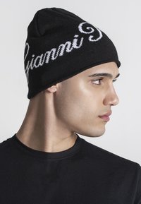 Zwarte gebreide beanie met witte cursieve "Gianni" tekst. Aansluitend ontwerp met een zachte textuur, gedragen boven een zwart shirt.