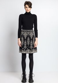 Crna rebrasta turtleneck majica, uparenjena s crnom suknjom do koljena koja ima bijele geometrijske uzorke i pojas. Uključuje crne vojne čizme.