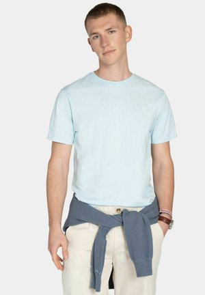 Joven con camiseta azul clara, pantalones beige y un suéter azul atado a la cintura, con un reloj y una pulsera en la muñeca.