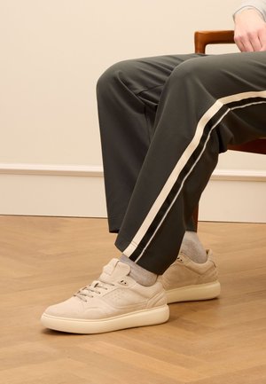 Beige suede sneakers with textured panels, lace-up design, and perforated accents. Worn with gray pants featuring side stripes.