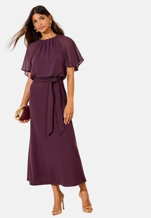 Femme portant une robe midi violette avec des manches papillonnantes, une taille nouée, tenant une pochette dorée et violette, et portant des sandales dorées à talons hauts.
