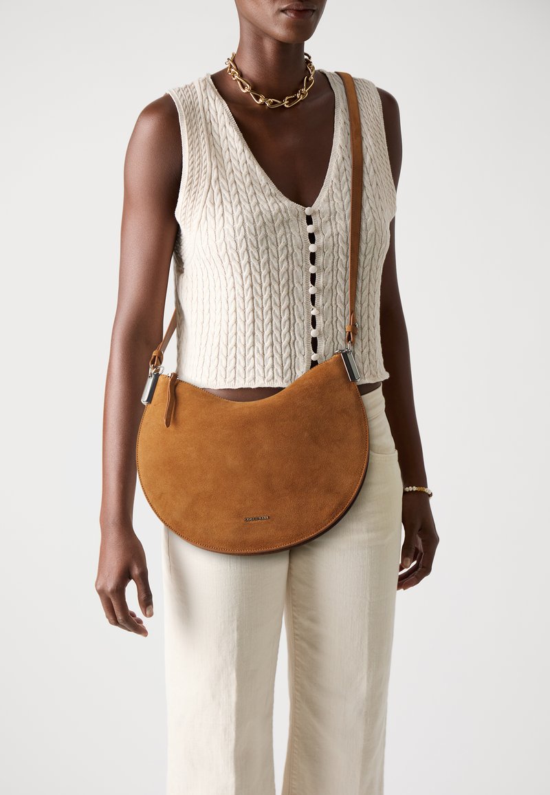 Borsa a tracolla in suede marrone a forma di mezzaluna, con accessori in argento e spallaccio. Abbinata a un top in maglia beige e pantaloni crema.