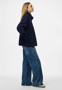 Marinblå fleecejacka med hög krage och fickor, parat med vida blå denimjeans och beige skor. Texturerad tygets detaljer synliga.