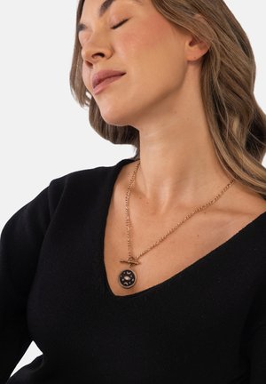 Frau mit geschlossenen Augen trägt ein schwarzes V-Ausschnitt-Oberteil und eine goldene Kette mit rundem schwarzem Anhänger, der ein Auge und Mondsymbole zeigt.