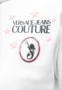 Logo Versace Jeans Couture z czarnym tekstem, różowymi gwiazdami i czarną syreną trzymającą trójząb, umieszczone wewnątrz różowego owalu na białym materiale.