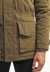 Gros plan d'une personne portant une veste Barbour marron avec des poches à rabat boutonnées et un poignet visible.