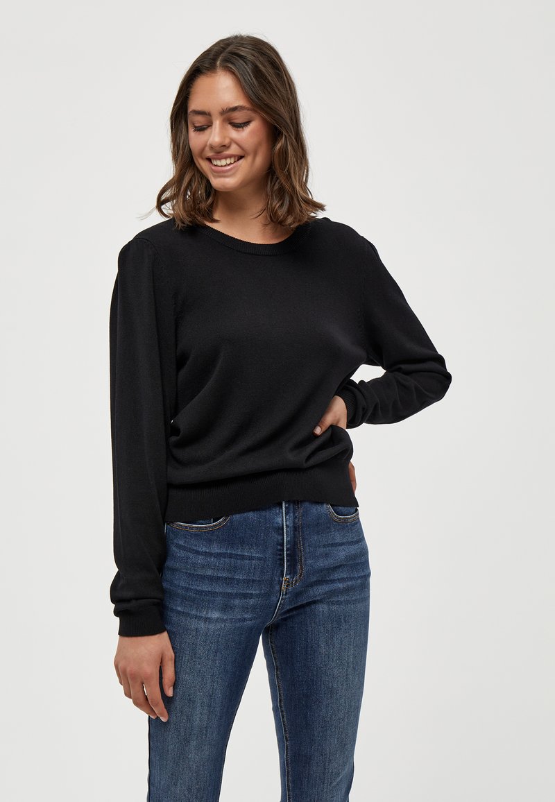 Maglione nero con maniche lunghe, scollatura a girocollo, polsini e orlo a costine. Indossato con jeans blu, che presentano una vestibilità ampia e una texture morbida.