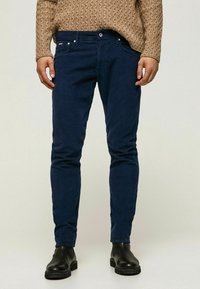 Man med slim-fit mörkblå kordflöjlsbyxor, tan kabelstickad tröja och svarta läderstövlar som står på en enfärgad ljus bakgrund.