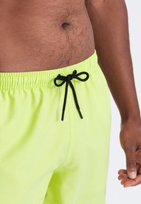 Neon keltainen uima-asu, jossa on joustava vyötärö ja musta naru. Kangas näyttää sileältä ja kevyeltä.