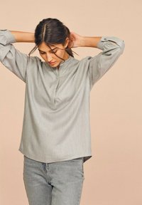 Lys grå langærmet skjorte med en mandarin krave, lavet af glat stof, der har et subtilt fald og opkrøllede ærmekanter, parret med lys grå jeans.