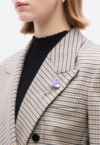 Μπεζ και καφέ σακάκι με μοτίβο καρό houndstooth και μαύρο ριμπ πλεκτό πουλόβερ από κάτω. Στο πέτο έχει στερεωθεί μια λιλά καρφίτσα σε σχήμα λουλουδιού με λεπτομέρειες από ασημένια καρφίτσα.