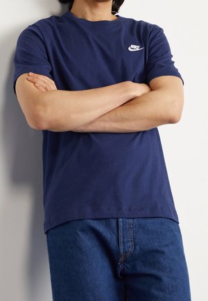 Personne portant un t-shirt Nike bleu marine, les bras croisés, debout devant un mur blanc uni, visible du cou jusqu'aux hanches.