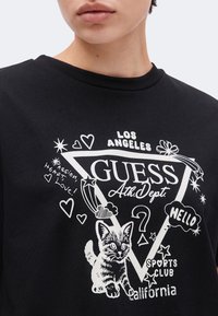 Personne portant un t-shirt noir avec un graphisme blanc de chat, cœurs, étoiles et texte incluant Guess, Los Angeles, Californie, Hello, et Sports Club.