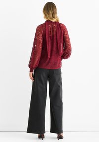 Burgundy sheer blouse with long sleeves and a tie detail at the back, paired with wide-leg black jeans and heeled shoes.