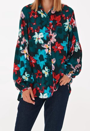 Femme portant une chemise ample à manches longues avec un grand imprimé floral rouge, bleu, blanc et rose sur tissu vert foncé, associée à un pantalon foncé.