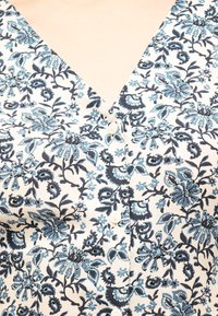 Blouse blanche avec motif floral bleu et noir, encolure en V et petits boutons blancs sur le devant.