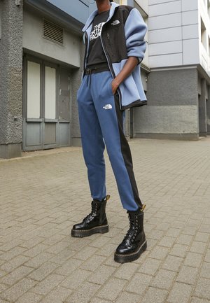 Person iført blå og sort The North Face træningsdragt med sorte platform snørestøvler stående på en brolagt gade.