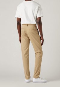 Chino in cotone beige con una vestibilità slim, caratterizzati da tasche posteriori e un piccolo etichetta sulla cintura, abbinati a una t-shirt bianca e sneakers bianche.
