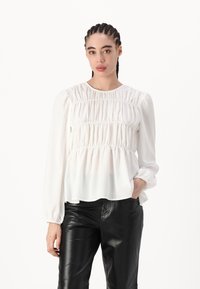 Bílá blůza s dlouhým rukávem a nabíranou texturou po celé látce, měkký materiál a mírný peplum střih, kombinovaná s černými koženými kalhotami.