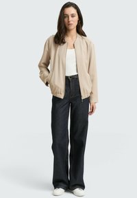 Veste bomber beige avec des poignets et un ourlet élastiques, portée sur un top blanc. Associée à un pantalon en denim large noir et des baskets blanches.