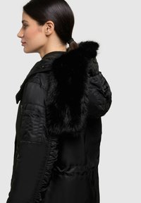 Melnā pūkainā jaka ar viltota pūka kapuci, ar rāvējslēdzēja kabatu, saburzītiem sānu šuvēm un gludu, spīdīgu audumu.