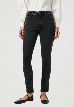Femme portant un jean noir slim, une chemise blanche à manches longues boutonnée, et des chaussures Mary Jane noires, debout devant un fond uni.