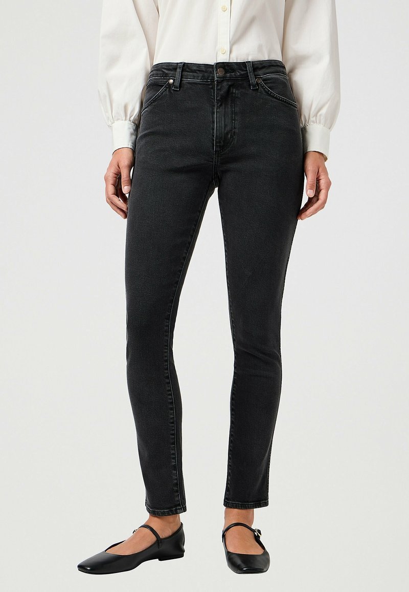 Femme portant un jean noir slim, une chemise blanche à manches longues boutonnée, et des chaussures Mary Jane noires, debout devant un fond uni.