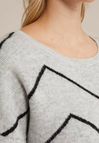 Šviesiai pilkas megztinis su minkšta tekstūra, papuoštas juodais zigzaginiais raštais. Platus iškirpimas ir suglebę rankogaliai praturtina dizainą.