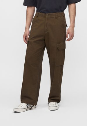 Person iført brune cargobukser med sidelommer og hvide casual sneakers, stående mod en ensfarvet baggrund.